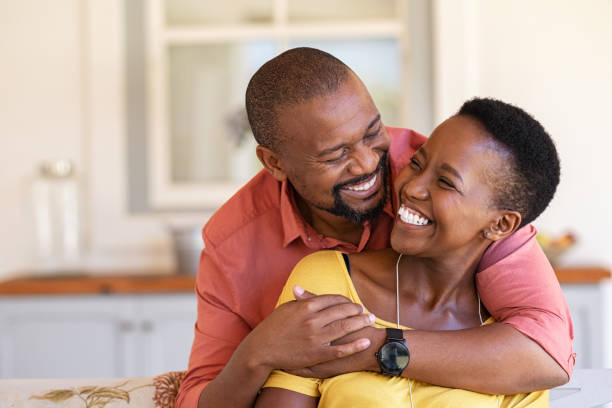 Mature black couple embracing on sofa while looking to each other. Romantic black man embracing woman from behind while laughing together. Happy african wife and husband loving in perfect harmony.